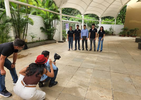 Photographers capturing a group photo of five people under a covered outdoor area with greenery in the background.
